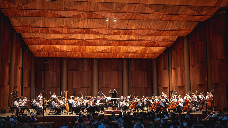 Wide view of an orchestra performing on a stage outdoors. 